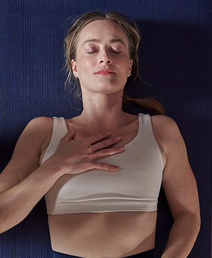 A woman lying down with her eyes closed, wearing a white ribbed tank top, with her hand resting on her chest against a dark blue Manduka yoga mat