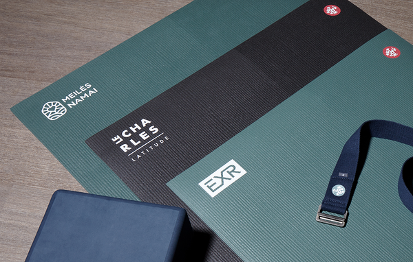 Manduka yoga mats in teal and black arranged on a wood floor with a yoga strap and dark foam blocks