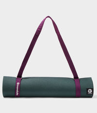 Porte-tapis de yoga Manduka maintenant un tapis de yoga vert foncé roulé à l’aide d’une bandoulière bordeaux sur un fond clair