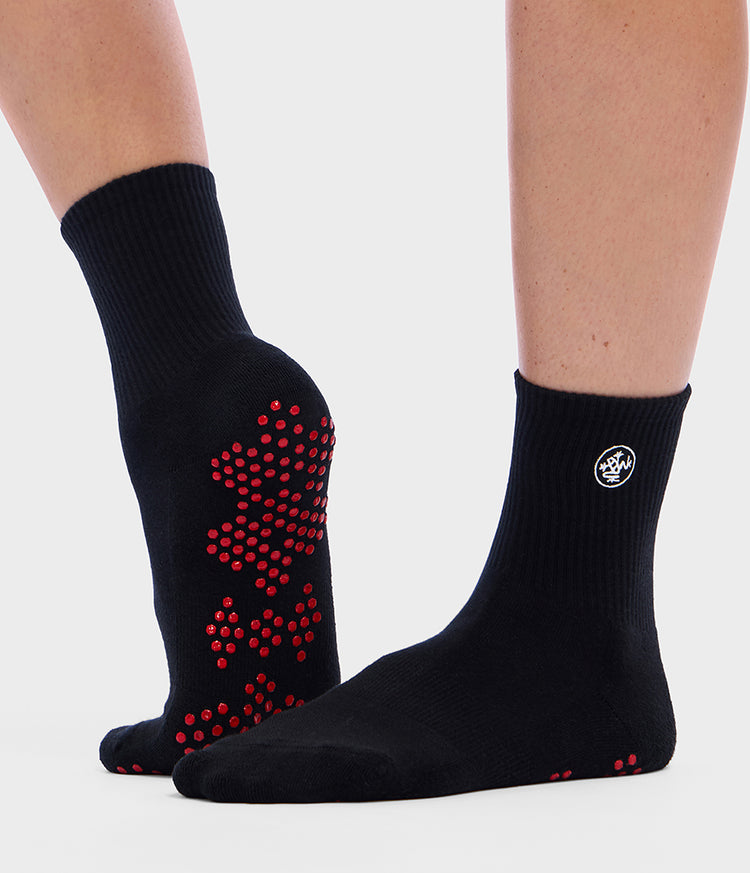 Chaussettes mi-mollet noires Manduka Equilibria pour le Pilates, portées aux pieds, avec des points d’adhérence en silicone rouge pour une meilleure stabilité et un placement précis des pieds pendant le Pilates sur reformer