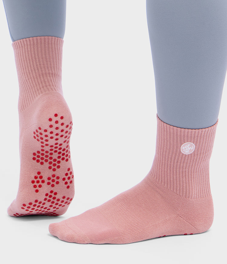 Chaussettes de Pilates roses Manduka à demi-mollet, portées aux pieds, avec des pastilles antidérapantes en silicone rouge sous un pied et une couverture côtelée à la cheville