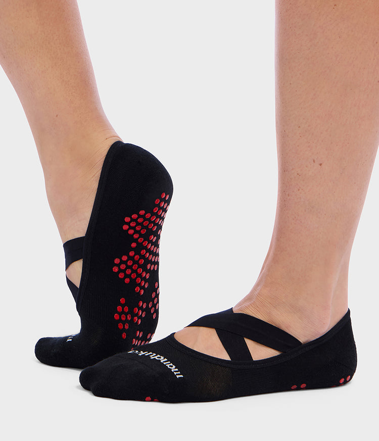 Manduka black Pilates slipper socks with red grip dots and crisscross straps shown on feet for stable reformer practice