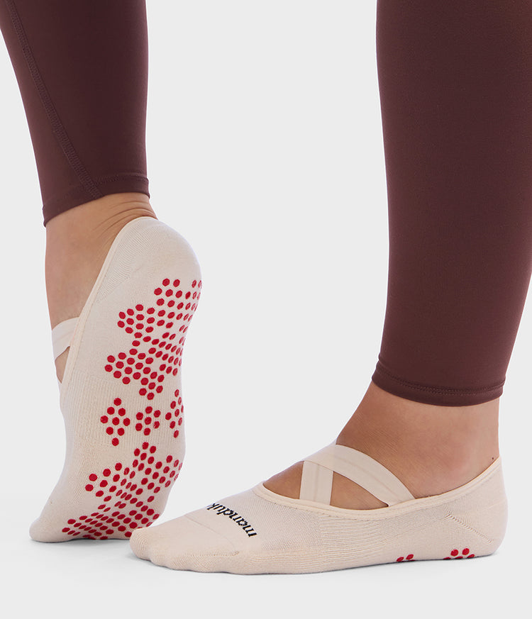 Manduka Pilates slipper socks in cream with soft elastic straps and a red silicone grip pattern shown on a person’s feet