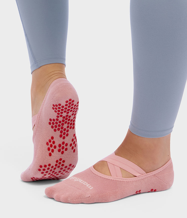 Manduka pink Pilates slipper socks with red silicone grip dots and ballet-inspired crossover straps shown on feet during reformer Pilates practice