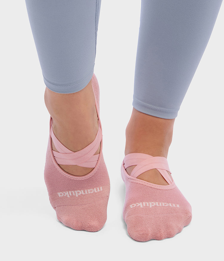 Manduka pink Pilates slipper socks with crossed elastic straps worn with gray leggings on a light background