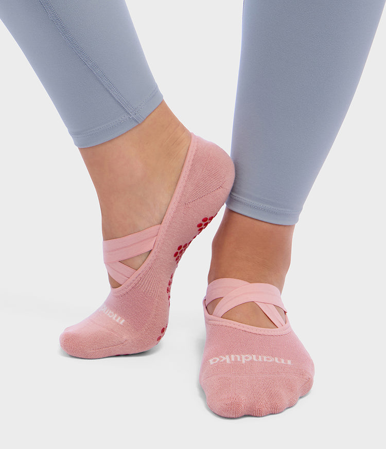 Manduka pink Pilates slipper socks with crossed elastic straps and red silicone grip pattern shown on a models feet