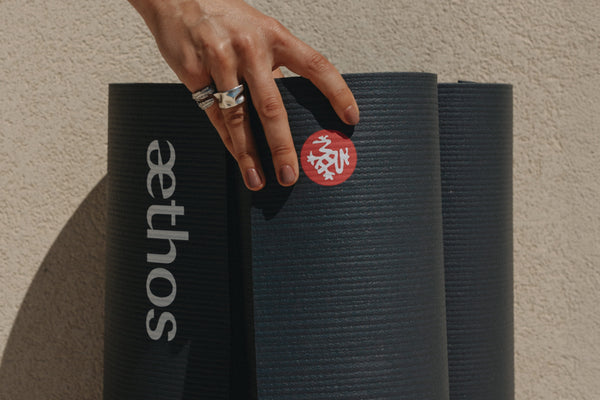 Hand with silver rings resting on a black textured yoga mat beside two other black mats with a red circular graphic and white ethos lettering