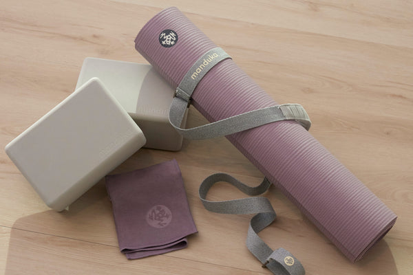 Rolled purple yoga mat with a gray carrying strap beside two white yoga blocks and a folded purple towel on a light wood floor