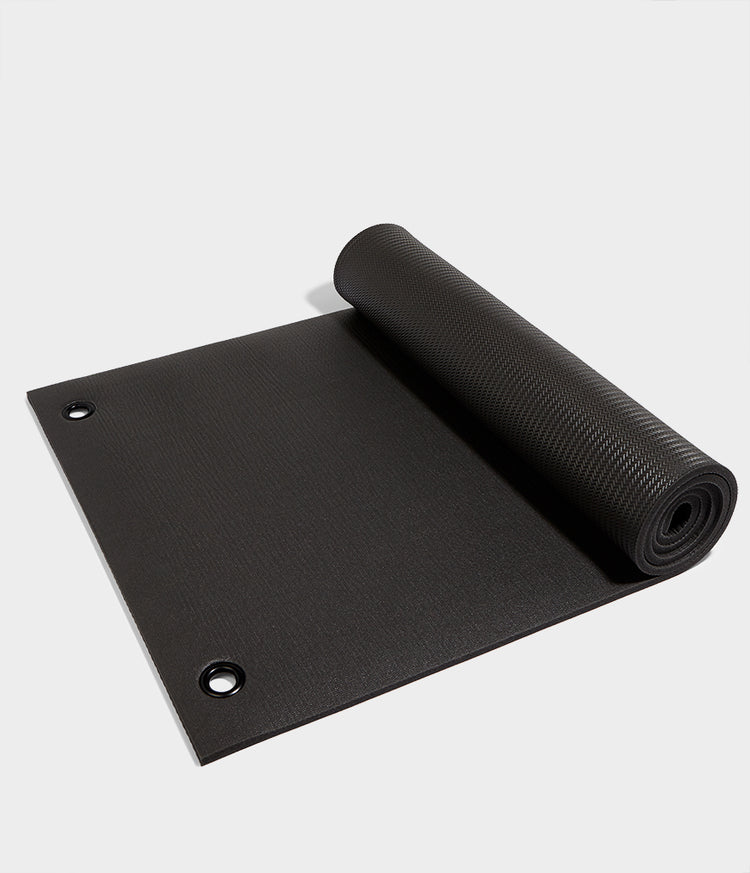 Manduka black fitness mat partially rolled out on a light background with a textured surface and reinforced corner grommets for gym workouts and floor exercises