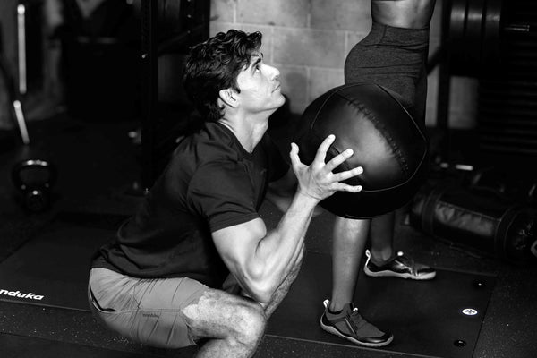 Black and white gym scene showing a man in a deep squat holding a large medicine ball while another person stands nearby on a Manduka exercise mat