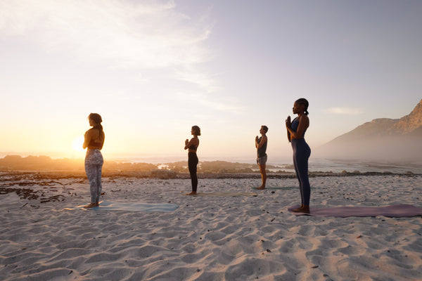 Four people practice yoga on a sandy beach at sunset with ocean waves and mountains in the background and a purple yoga mat in the foreground