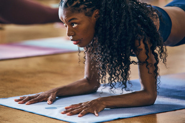Woman practicing a yoga pose on a blue Manduka yoga mat in a warm wood floored studio with another person blurred in the background