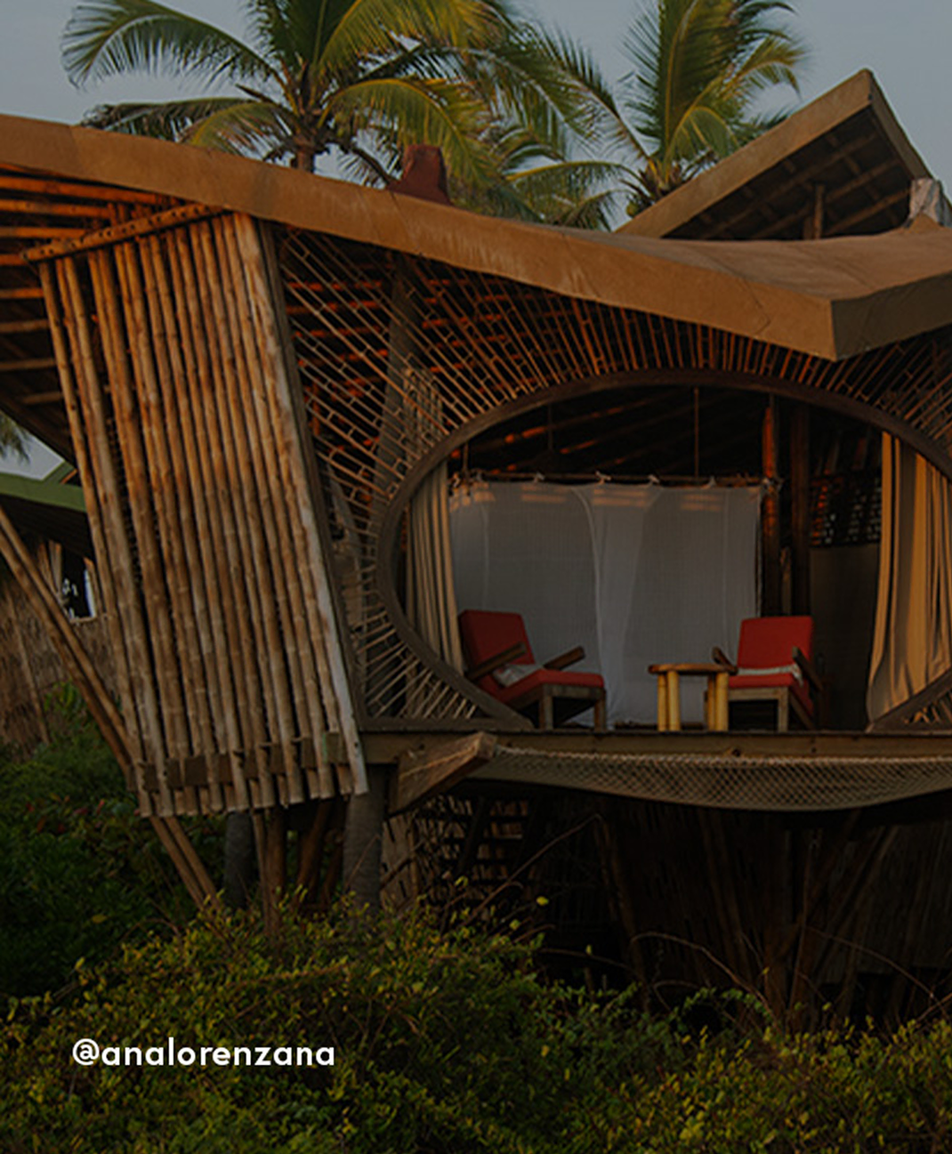 Elevated bamboo hut with curved woven walls, red lounge chairs, and a small table surrounded by tropical greenery and palm trees
