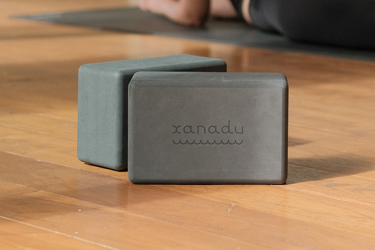 Two gray Manduka yoga blocks with a xanadu logo resting on a wooden studio floor beside a person practicing yoga