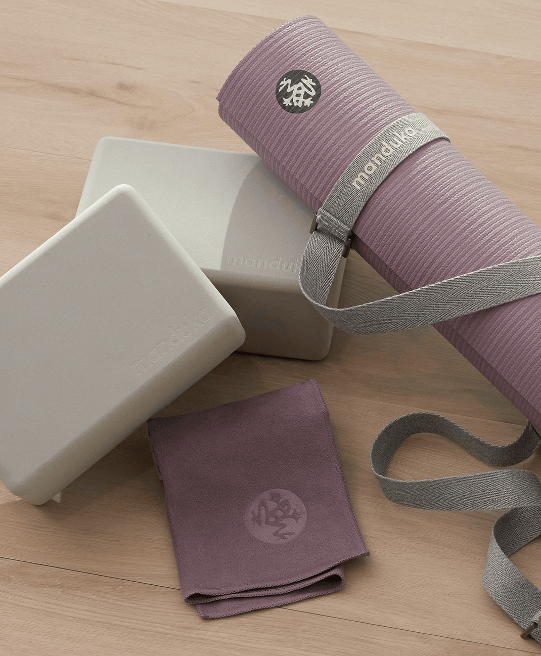 Rolled mauve Manduka yoga mat with carrying strap beside two cream yoga blocks and a folded purple towel on a wooden floor