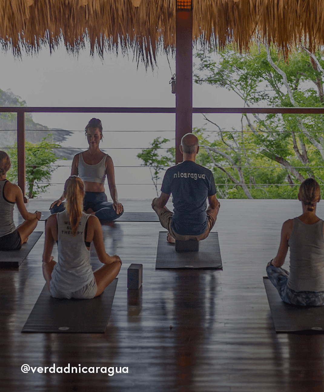 Group of people practicing yoga on mats in an open tropical pavilion with thatched roofing, surrounded by trees and mountain views.