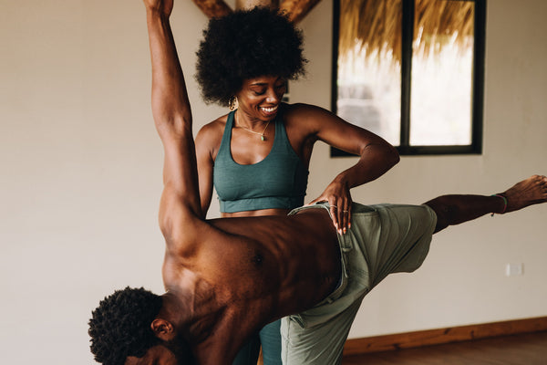 Two people practice an acrobatic yoga pose indoors with the woman supporting a man as he balances horizontally with one leg extended