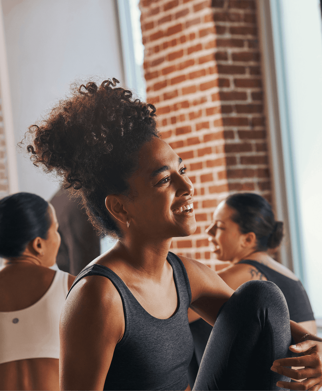 Woman in a black workout top smiles during a yoga class with one knee raised as other participants stretch in a bright brick-walled studio