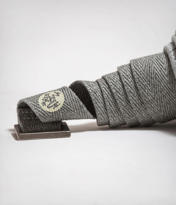 Close up of a gray Manduka mat carrier strap with textured fabric wrapped beside a metal buckle and branded detail